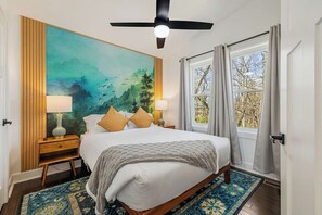 A bedroom with a bed, two bedside tables, a ceiling fan, a large window with curtains, and a wall mural.