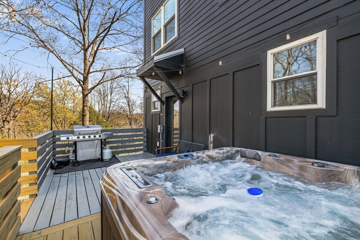 A hot tub on a wooden deck with a grill and a portable heater, adjacent to a house with dark siding and windows.