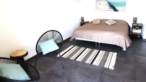 Une chambre à coucher comprenant un lit, deux tables de chevet, une chaise, une petite table, un tapis à rayures et un tableau accroché au mur représentant une jetée.