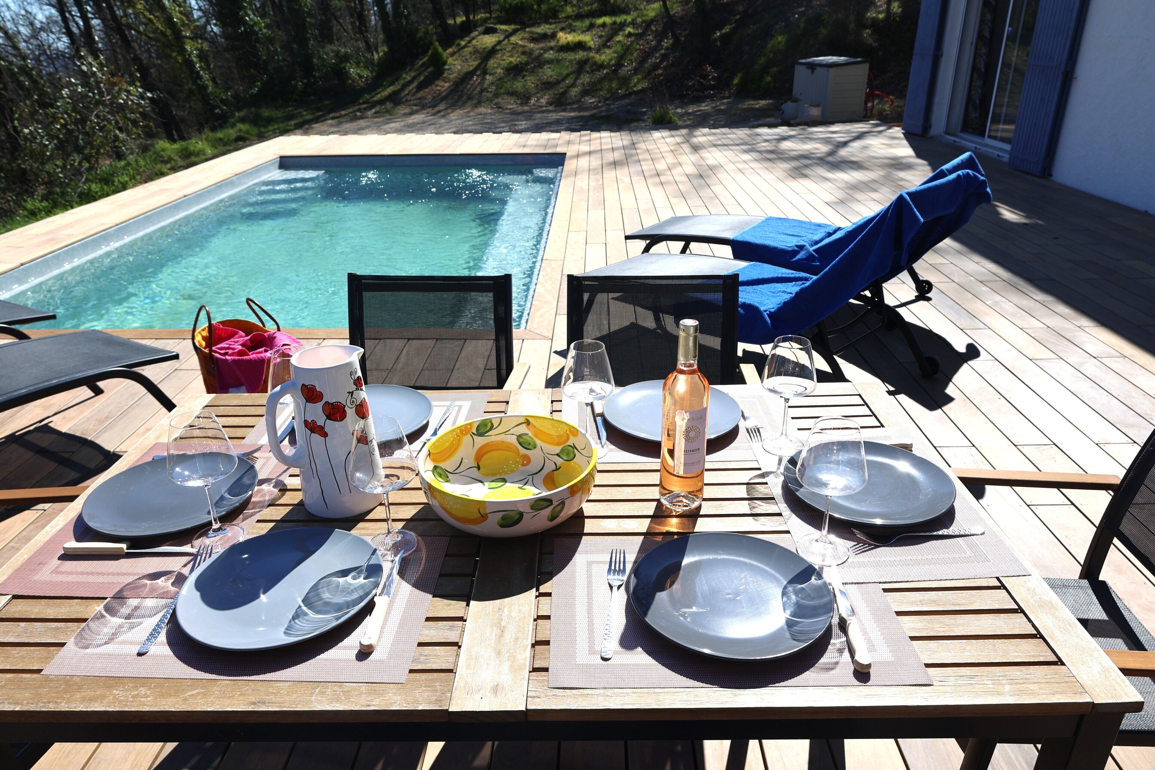 A wooden deck with a pool, dining table set with plates, glasses, and a bottle.