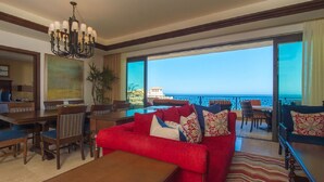 A hotel room with a red sofa, a wooden coffee table, a dining area with chairs, and a large window offering a view of the ocean. - Spacious 2 BR, 3 bath Presidential Suite. Pacific Ocean View. 6 guests welcome! (Cabo San Lucas)