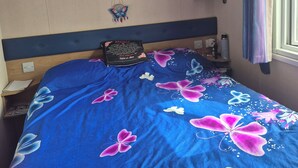 Un dormitorio con una cama, una mesita de noche, un estante con ropa doblada, un estante en la pared con una cesta y una ventana con cortinas.