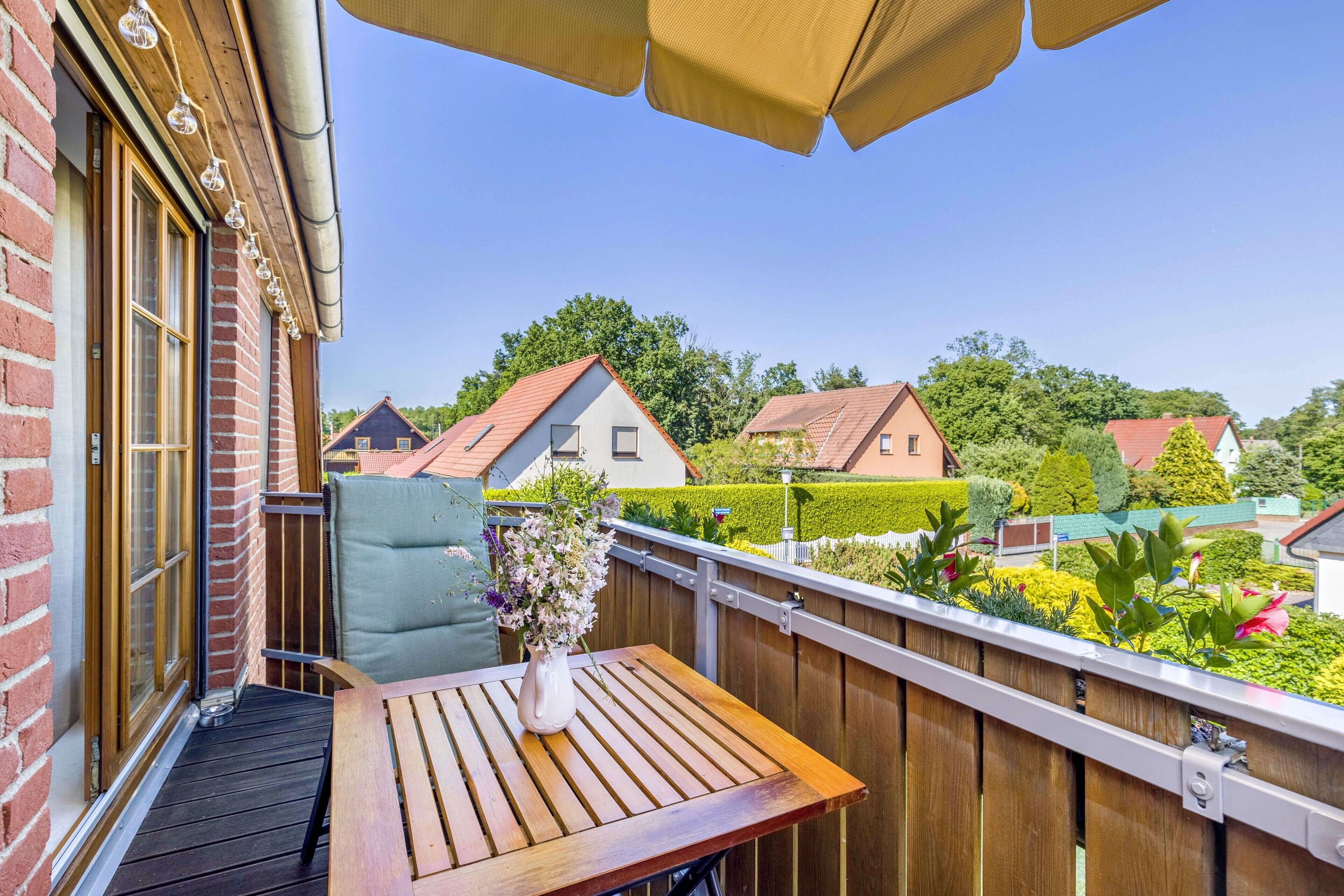 A wooden deck with a table and a chair, a potted plant, and a view of houses and greenery in the background.