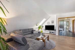 Un salon moderne avec un canapé d’angle gris, une table basse en bois, un téléviseur à écran plat et une grande fenêtre.