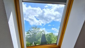 Une fenêtre de toit donnant sur les arbres et le ciel, une lampe blanche posée sur une table de chevet, et une vue partielle d’un lit.