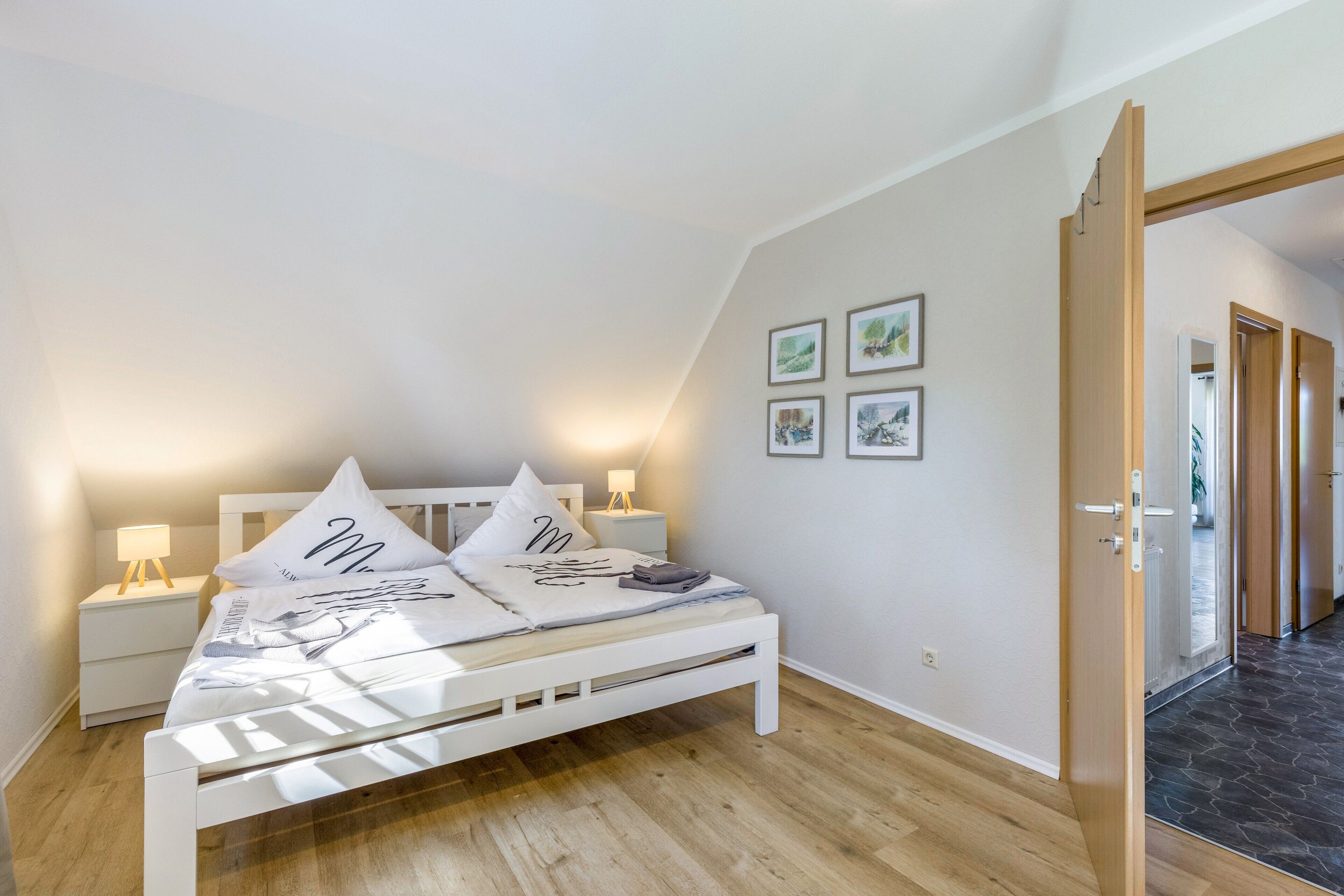Un dormitorio con cama, mesitas de noche, decoración en la pared y una puerta que da a otra habitación.