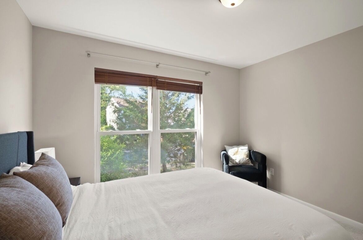 A bedroom with a large bed, a chair, a window with blinds, and a ceiling light.