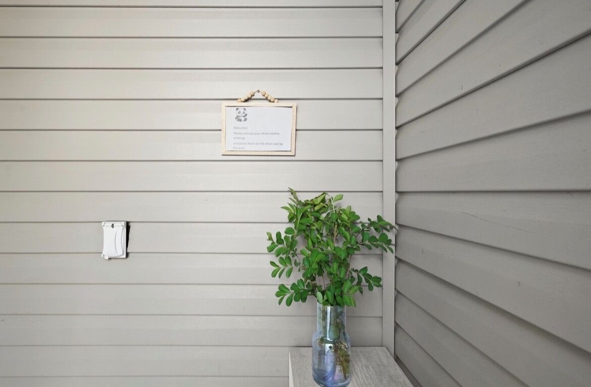 Un letrero en la pared con un mensaje de bienvenida, una pequeña caja eléctrica blanca y un florero de vidrio transparente con una planta verde.