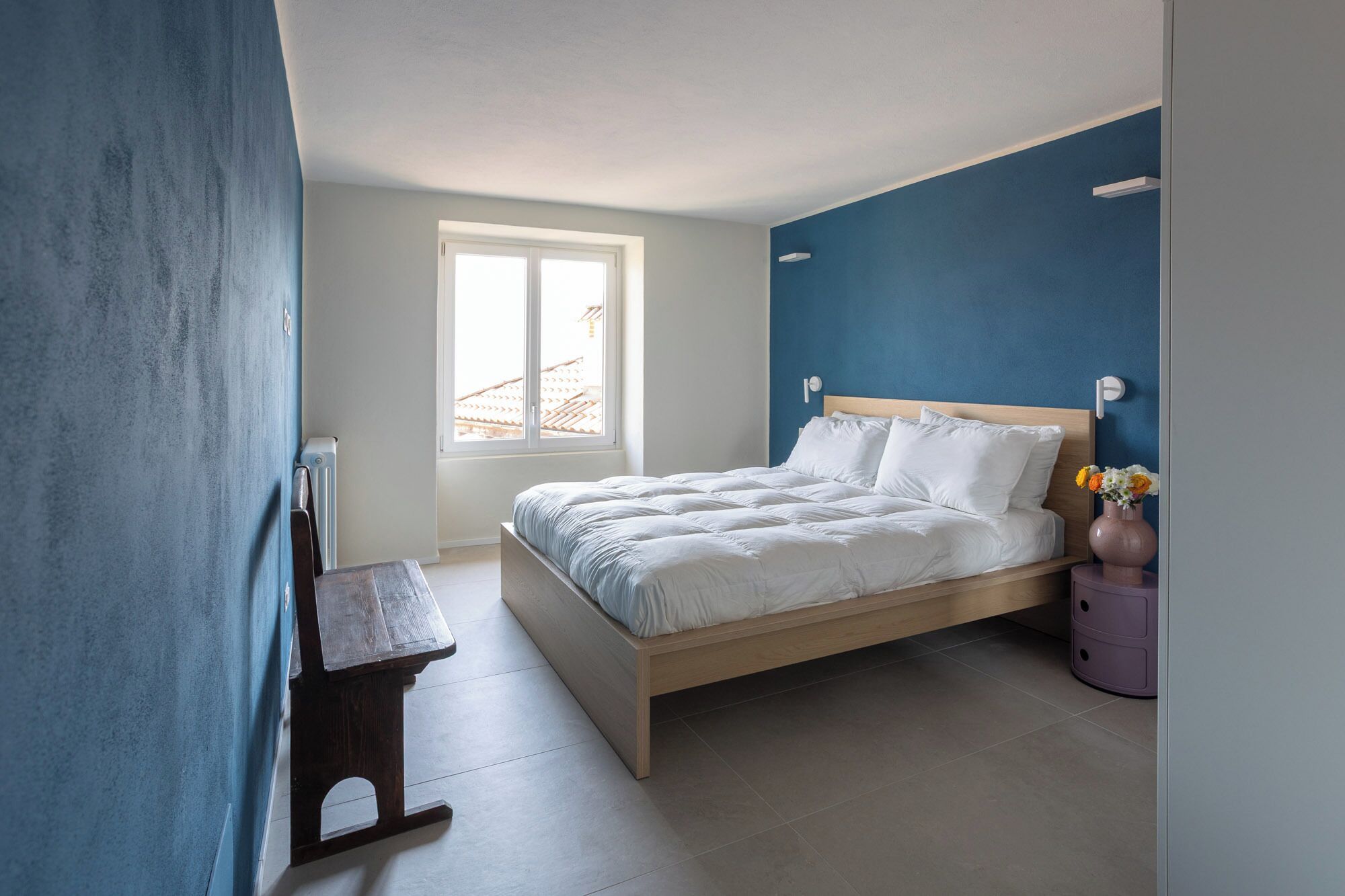 Un dormitorio con cama, ventana, radiador, mesita de noche y un florero con flores.