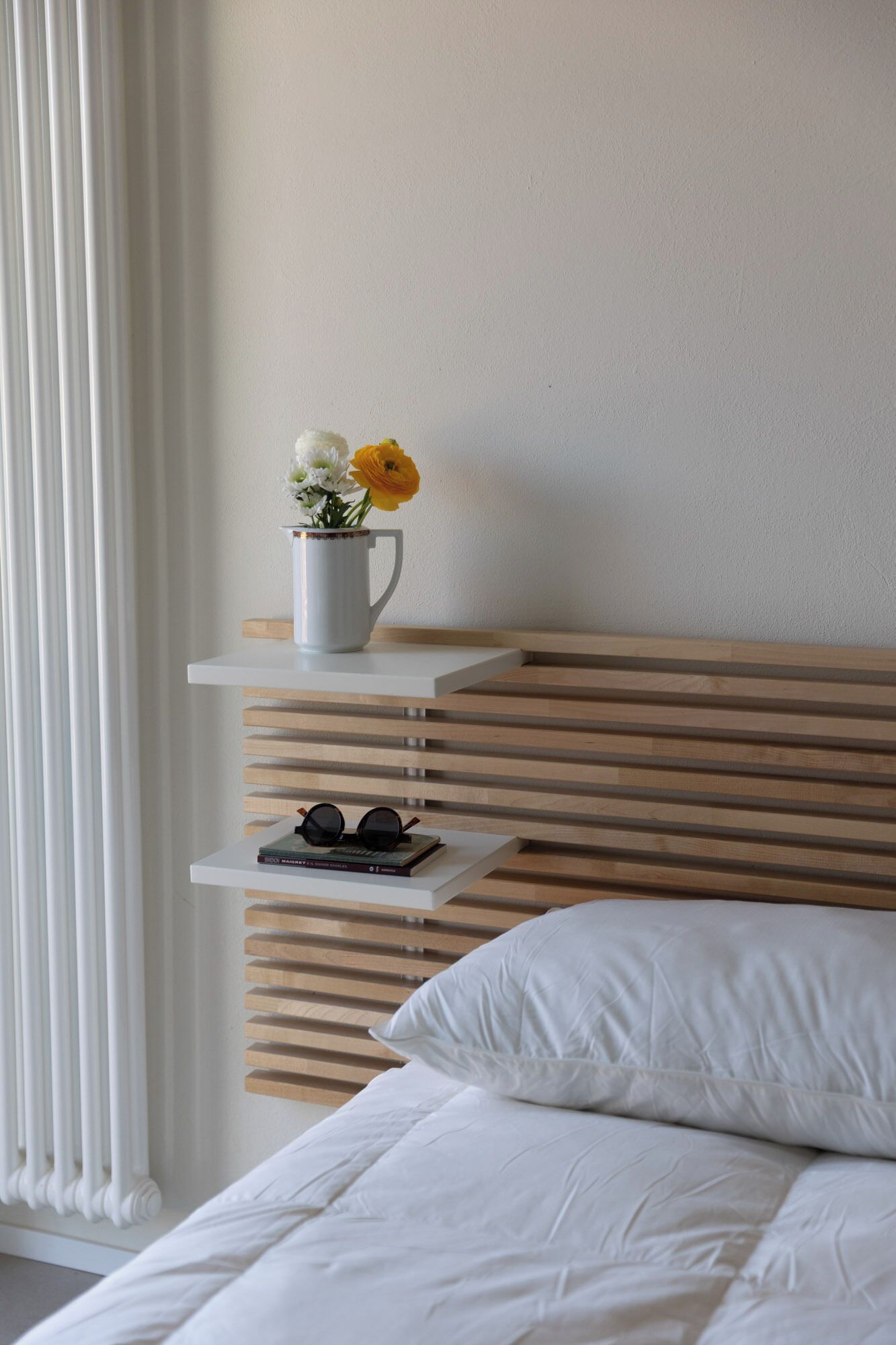 A bedroom with a bed, a white pillow, a white nightstand with a cup of flowers, and a pair of glasses.