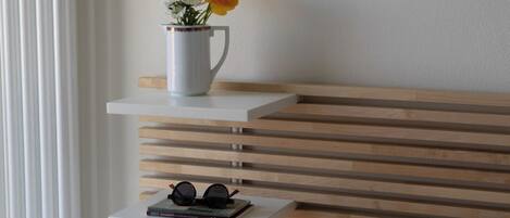 A bedroom with a bed, a white pillow, a white nightstand with a cup of flowers, and a pair of glasses.