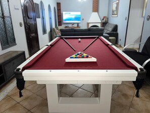 A pool table with a triangular rack of billiard balls, a television in the background, and a sofa.