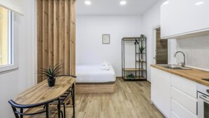 Une cuisine compacte avec un plan de travail en bois, une table à manger avec des chaises, un lit avec un matelas blanc et une petite étagère avec des plantes.
