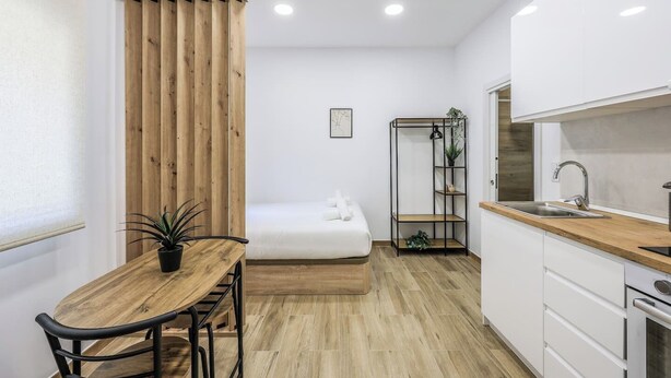 Une pièce de taille modeste comprenant un lit, une petite table à manger avec des chaises, un coin cuisine et un mur décoratif en bois.