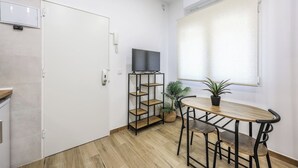 Une petite pièce moderne avec un plancher en bois, une porte blanche, un téléviseur posé sur un meuble noir, une table ronde avec deux chaises et une plante en pot.