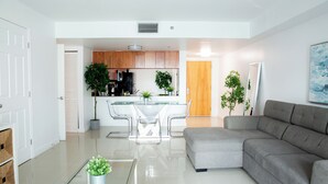 Ein modernes Wohnzimmer mit einem grauen Sofa, einem Glastisch und einer Küche im Hintergrund.