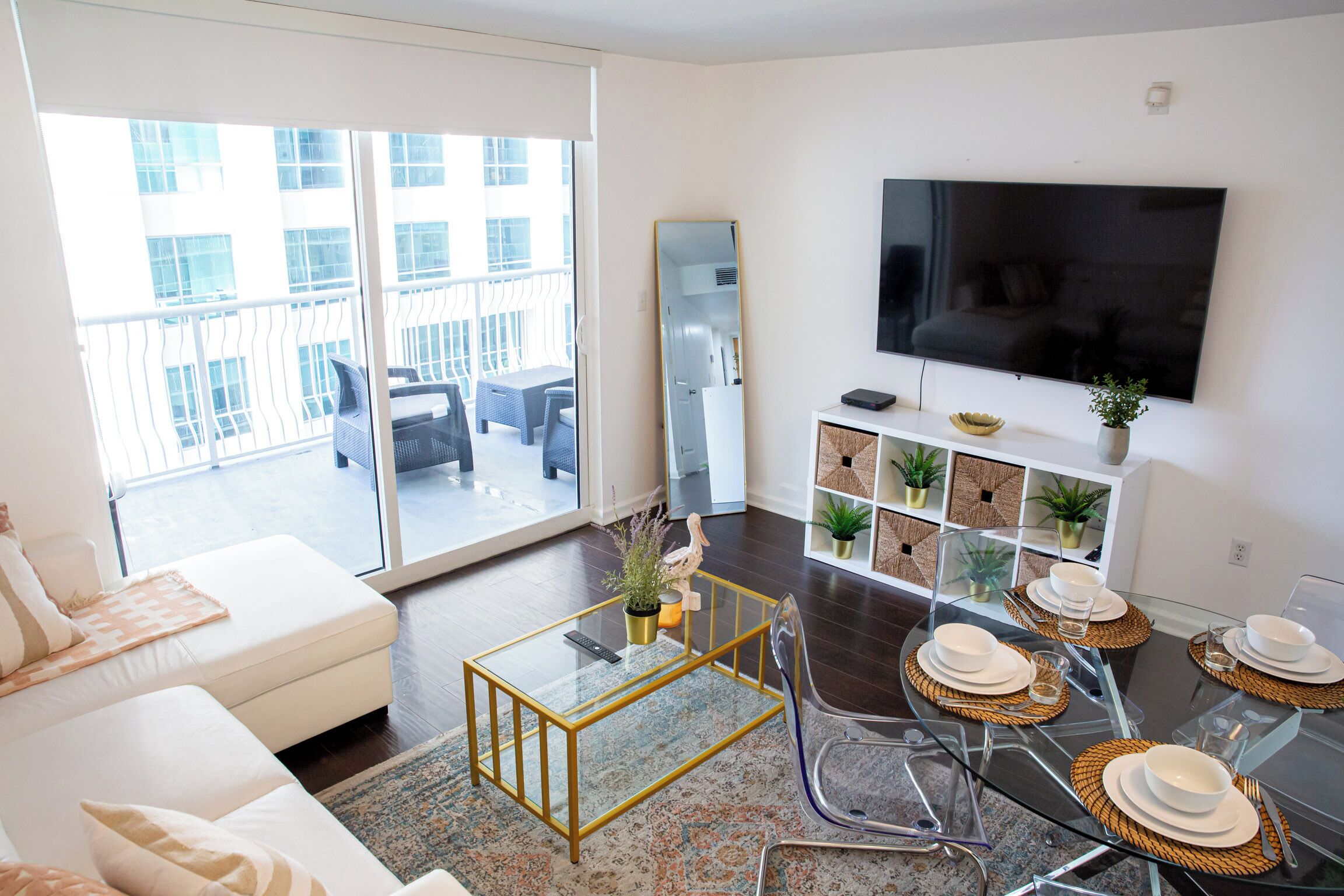 A modern living room with a glass coffee table, a sofa, a dining table set for four, and a large flat-screen TV.