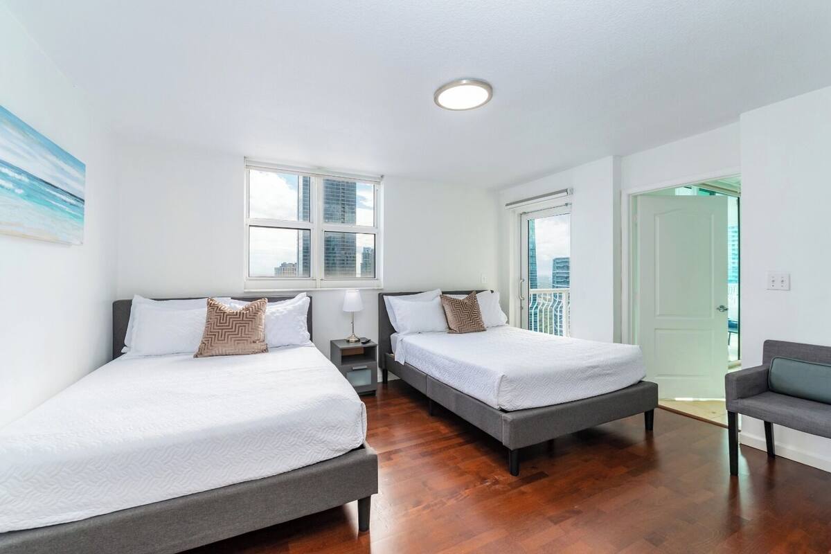 Quarto moderno com duas camas, uma janela, uma porta e um espaço de estar pequeno.