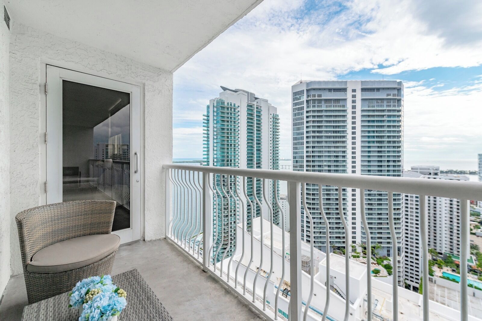 A balcony with a wicker chair and a vase of flowers, overlooking high-rise buildings.