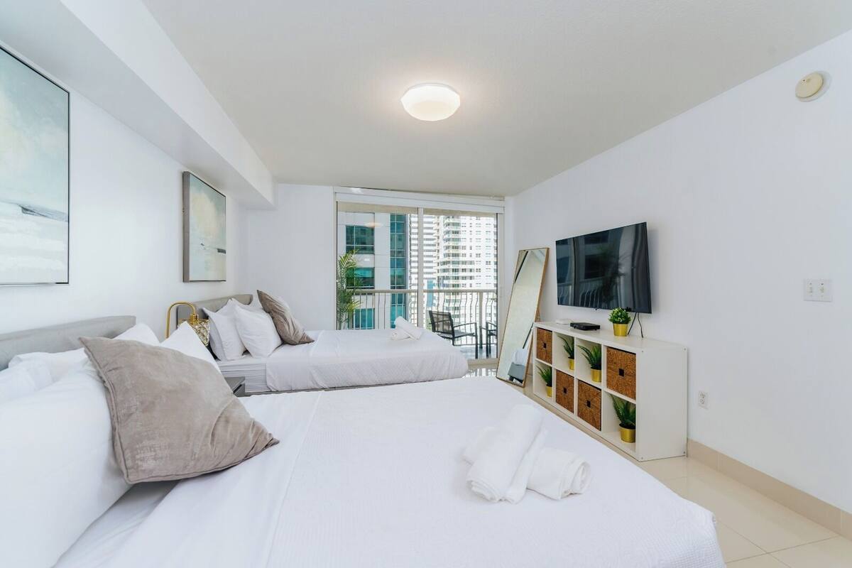 Quarto de hotel com duas camas, televisão, espelho e varanda com vista para a cidade.