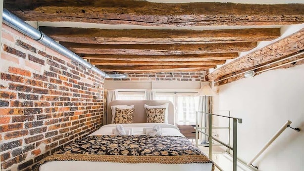 Une pièce étroite aux murs en briques apparentes, avec un lit recouvert d’une courtepointe à motifs et une balustrade en métal.