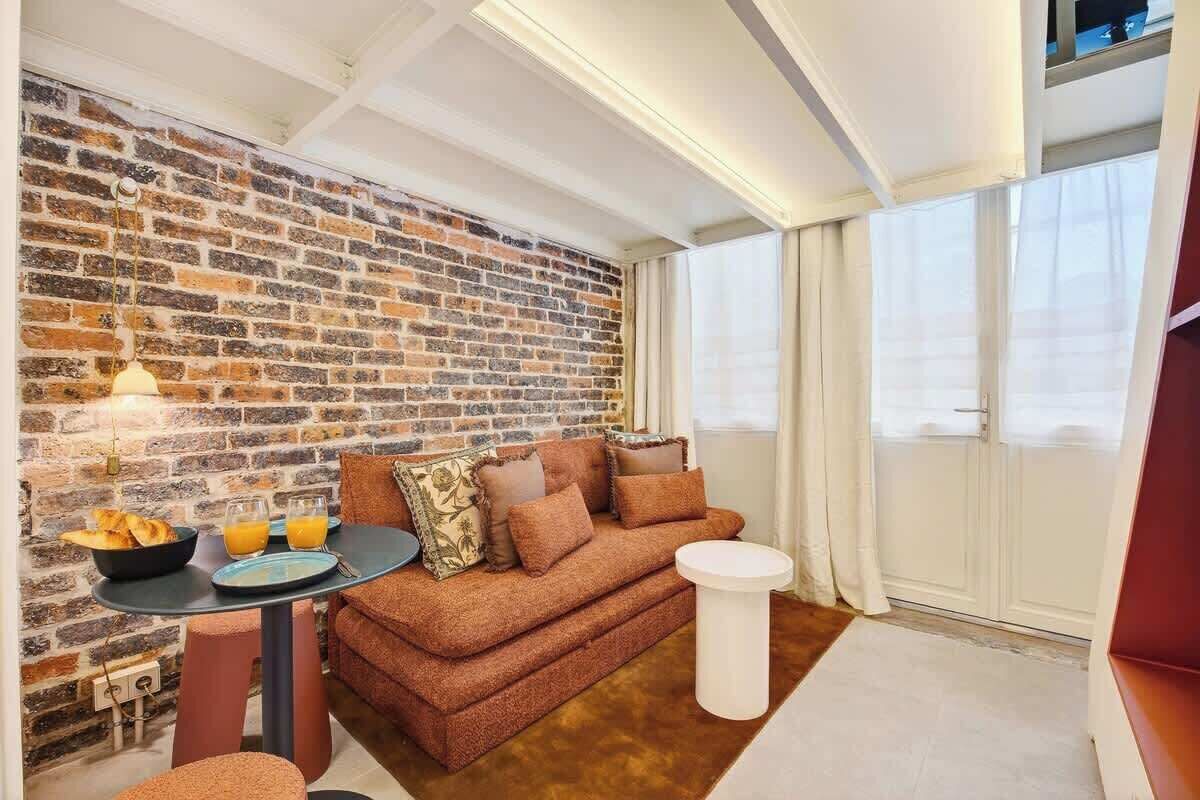 A cozy living area with a brick wall, a sofa, a round table with breakfast items, and a large window.