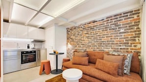 Un espace de vie compact comprenant un coin cuisine, un mur décoratif en briques, un canapé avec des coussins et une petite table ronde.