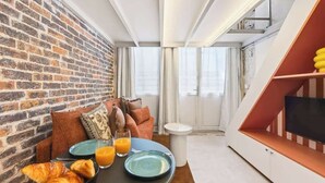 Une pièce moderne avec un mur en briques, un canapé, une table à manger avec des assiettes et des croissants, et un téléviseur posé sur une étagère.