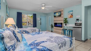 A room with a bed, a TV, a kitchenette, and a window with curtains. - Cozy 2 Queen Bed Studio in Sunny Fort Myers Beach (Fort Myers Beach)