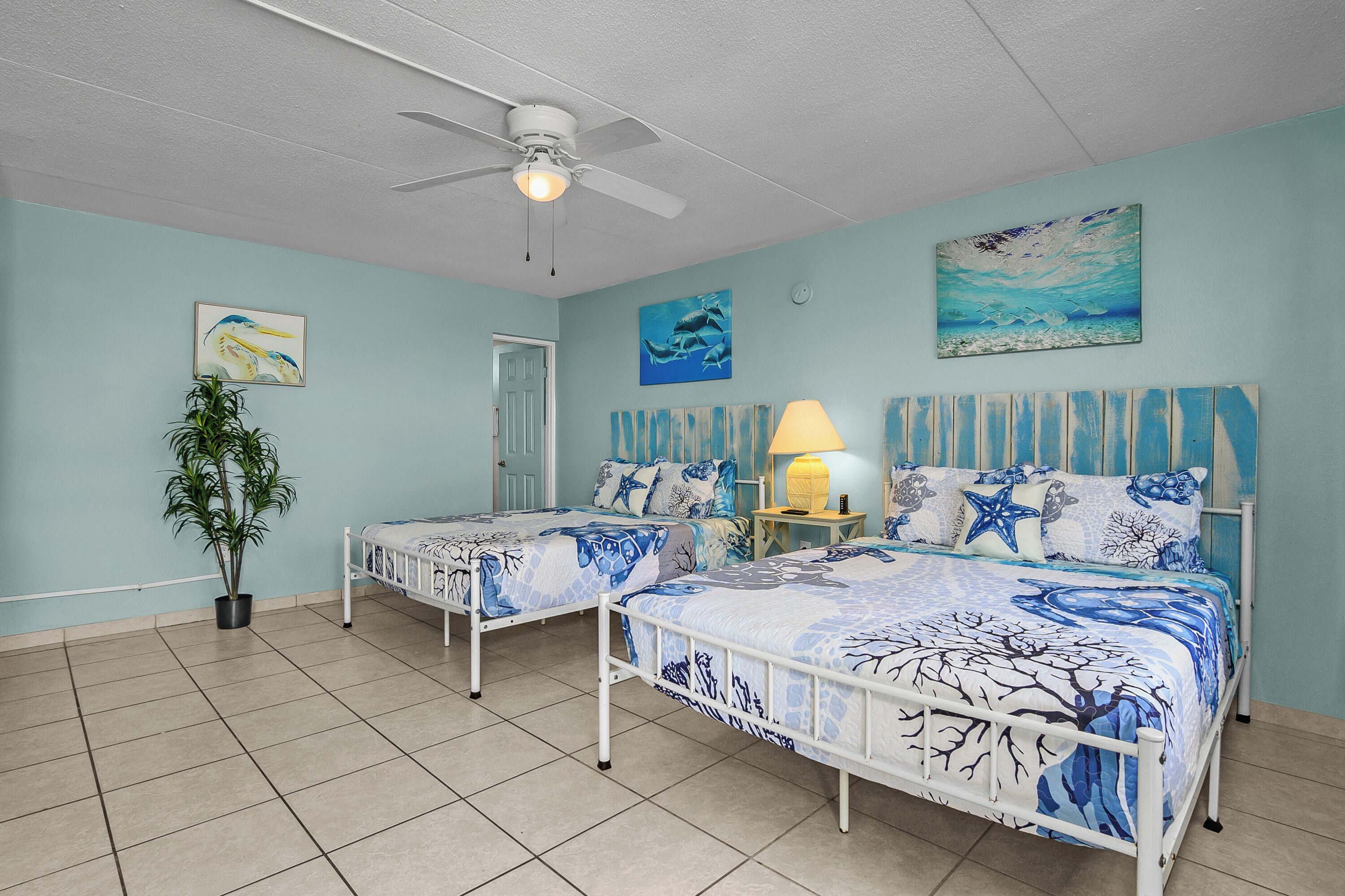 A bedroom with two beds, a ceiling fan, a potted plant, and artwork on the walls.