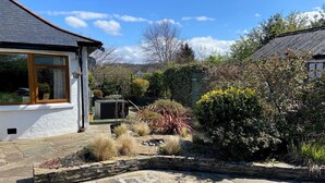 Un patio de piedra que conduce a una casa blanca con una ventana y un jardín con diversas plantas y un muro de piedra.