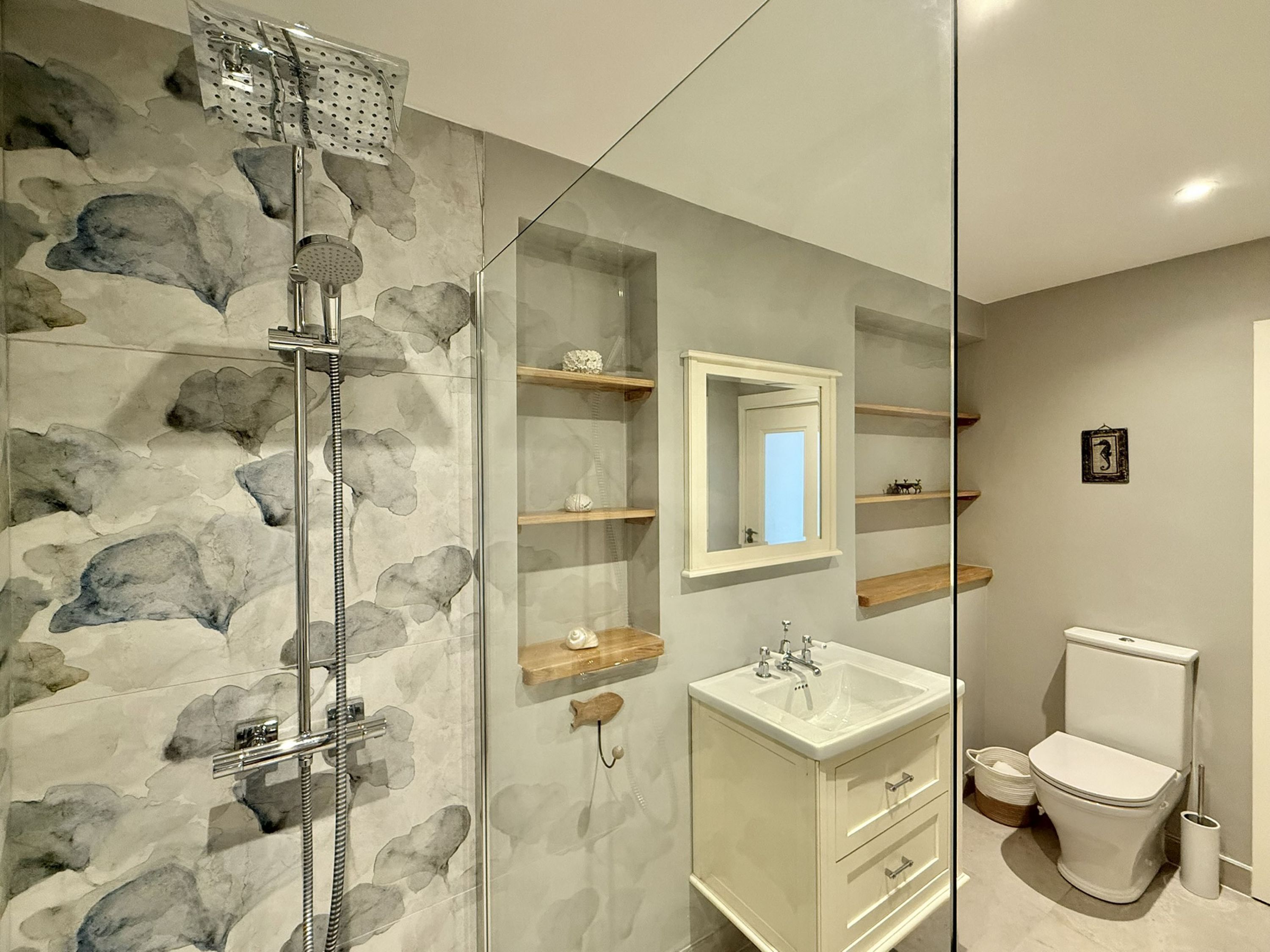 A bathroom with a glass shower enclosure, a white sink, a toilet, and wooden shelves.