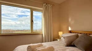 A hotel room with a large window, a bed with rolled towels, a nightstand with a lamp, and a view of the countryside. - Cottage 498 - Cleggan - sleeps 9 guests  in 4 bedrooms (Cleggan)