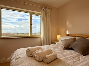 Ein Hotelzimmer mit großem Fenster, einem Bett mit zusammengerollten Handtüchern, einem Nachttisch mit Lampe und Blick auf die Landschaft.