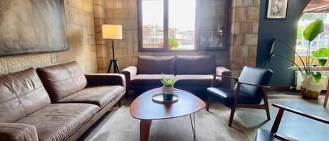 Una sala de estar con un sofá de cuero marrón, una mesa redonda con una planta, un sillón azul y una pequeña mesa con una silla.