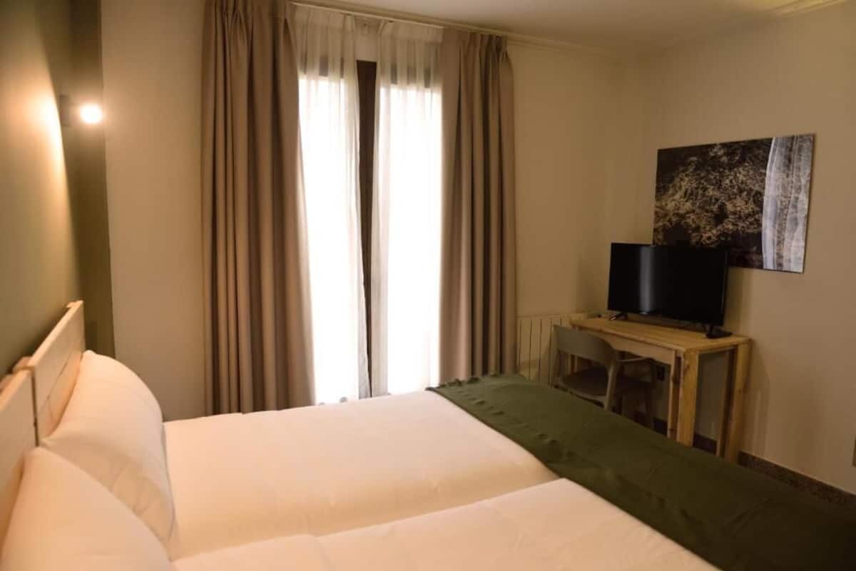 Habitación de hotel con cama, cortinas, un televisor sobre una mesa de madera y un cuadro en la pared.