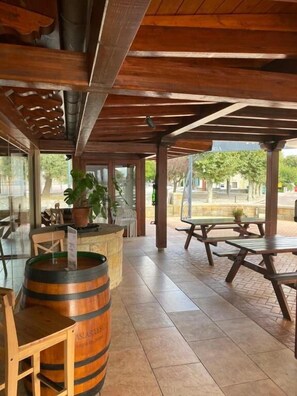 Een terras buiten met houten tafels en stoelen, een houten vat en een bloempot.