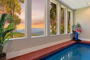 Une piscine intérieure avec un panier de basket, de grandes fenêtres offrant une vue sur les arbres et un coucher de soleil, et une plante sur la gauche.