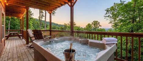A hot tub with champagne, glasses, and strawberries on a wooden deck with a view of trees.