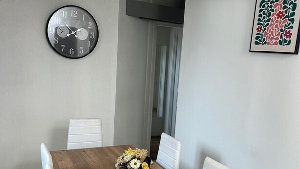 A dining area with a wooden table, white chairs, a clock, and a floral artwork on the wall.