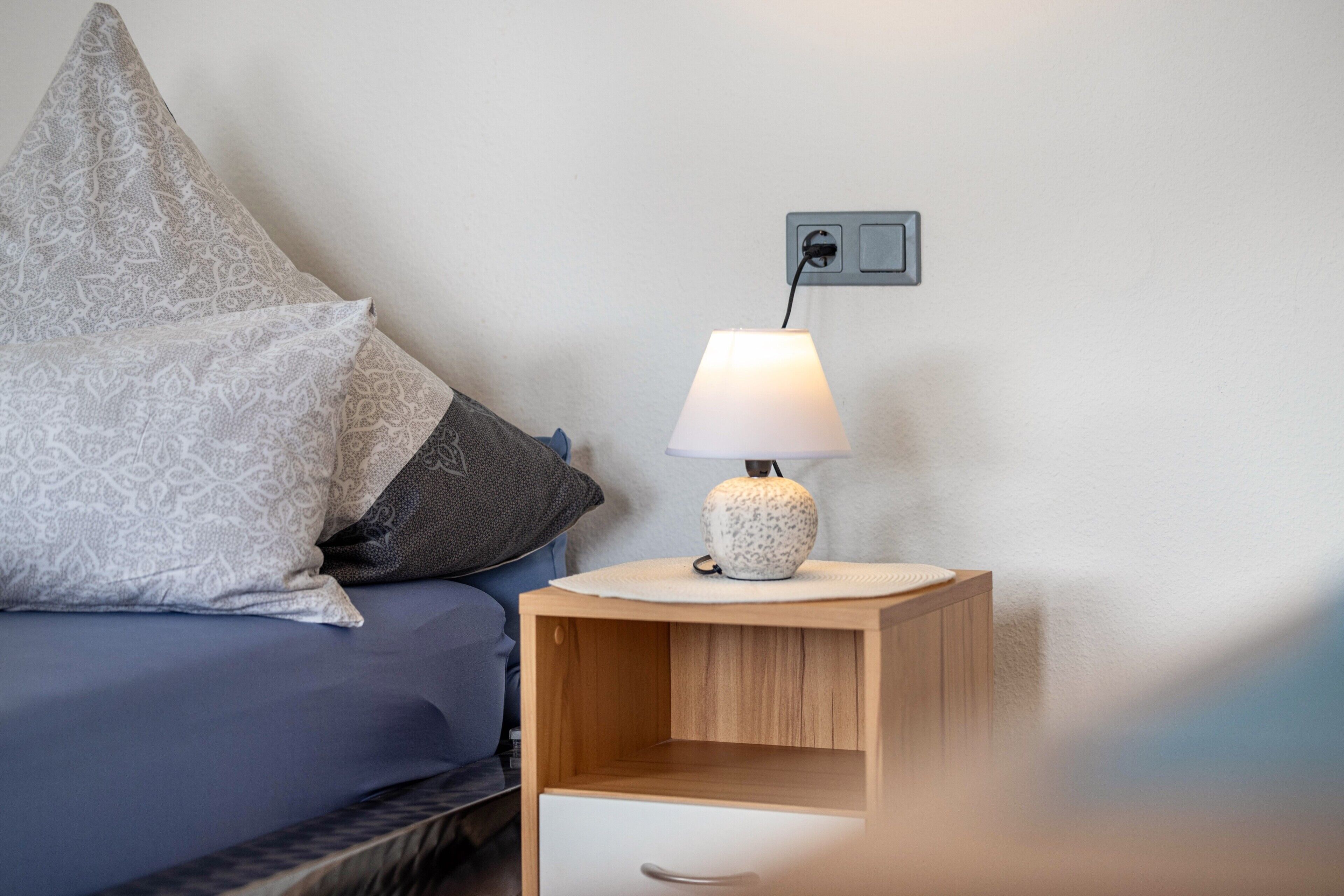A bedroom with a bed, pillows, a bedside table with a lamp, and a power outlet.