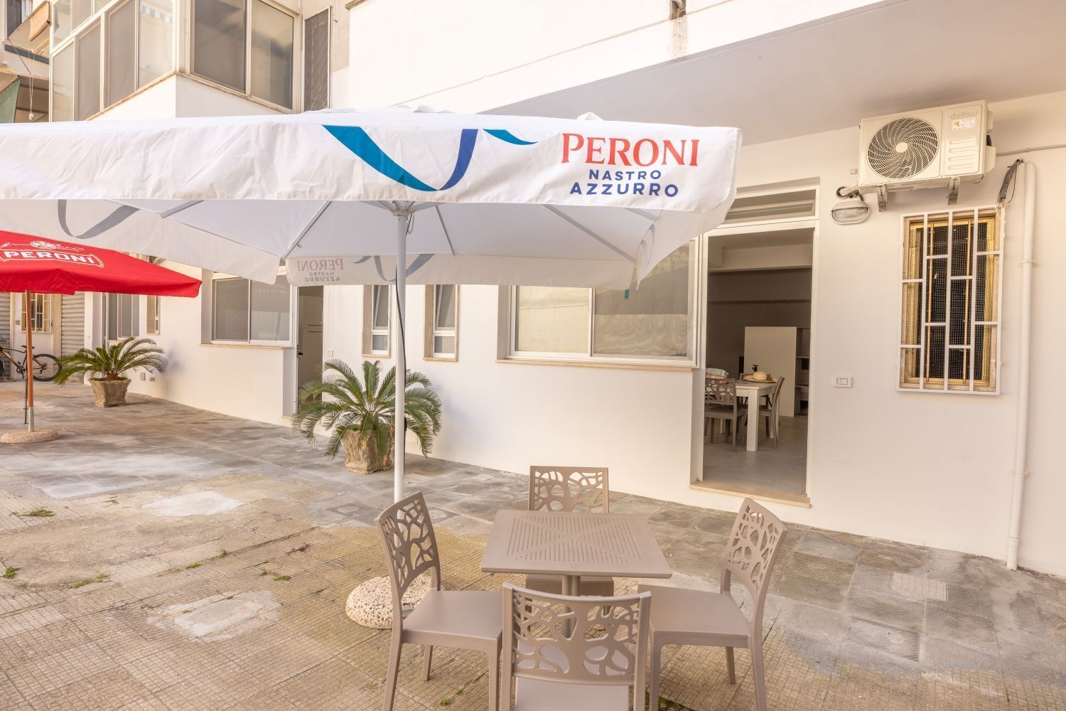 An outdoor seating area with a table and chairs, a red umbrella, and a building with a sign reading 'PERONI Nastro Azzurro.'