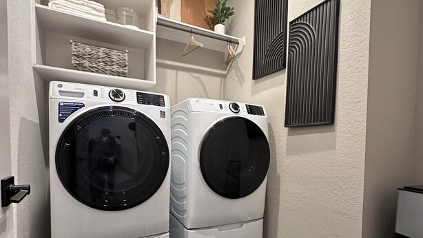Un espace buanderie comprenant deux machines à laver à chargement frontal superposées, une étagère avec des serviettes pliées, une plante suspendue et deux œuvres d'art abstraites accrochées au mur.