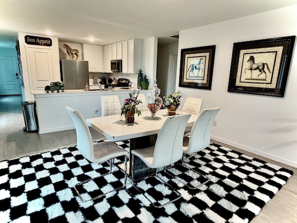 Una cocina moderna con armarios blancos, un refrigerador y un microondas. Hay un comedor con una mesa, sillas y un tapete a cuadros blanco y negro.