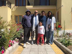 A family standing in front of a building with a balcony, a garden with flowers, and a paved walkway.