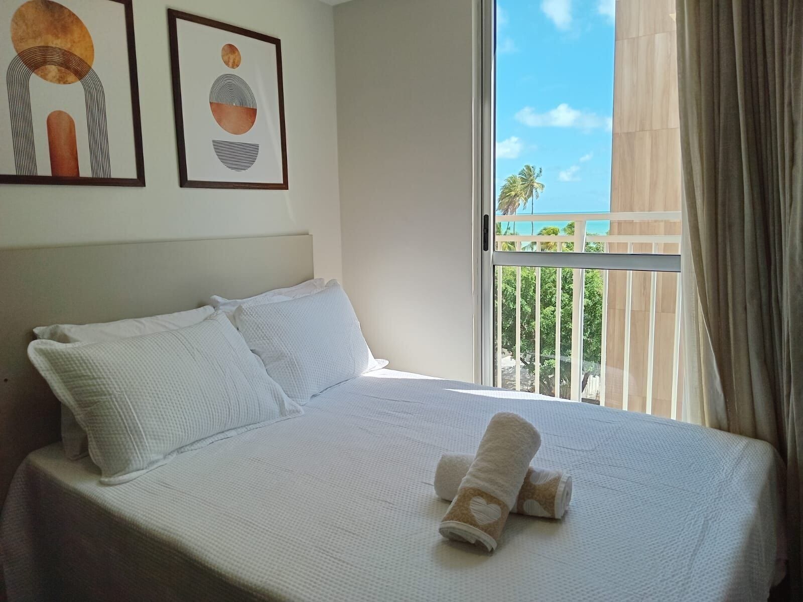 A hotel room with a bed, pillows, a window with a view of palm trees, and two framed artworks on the wall.