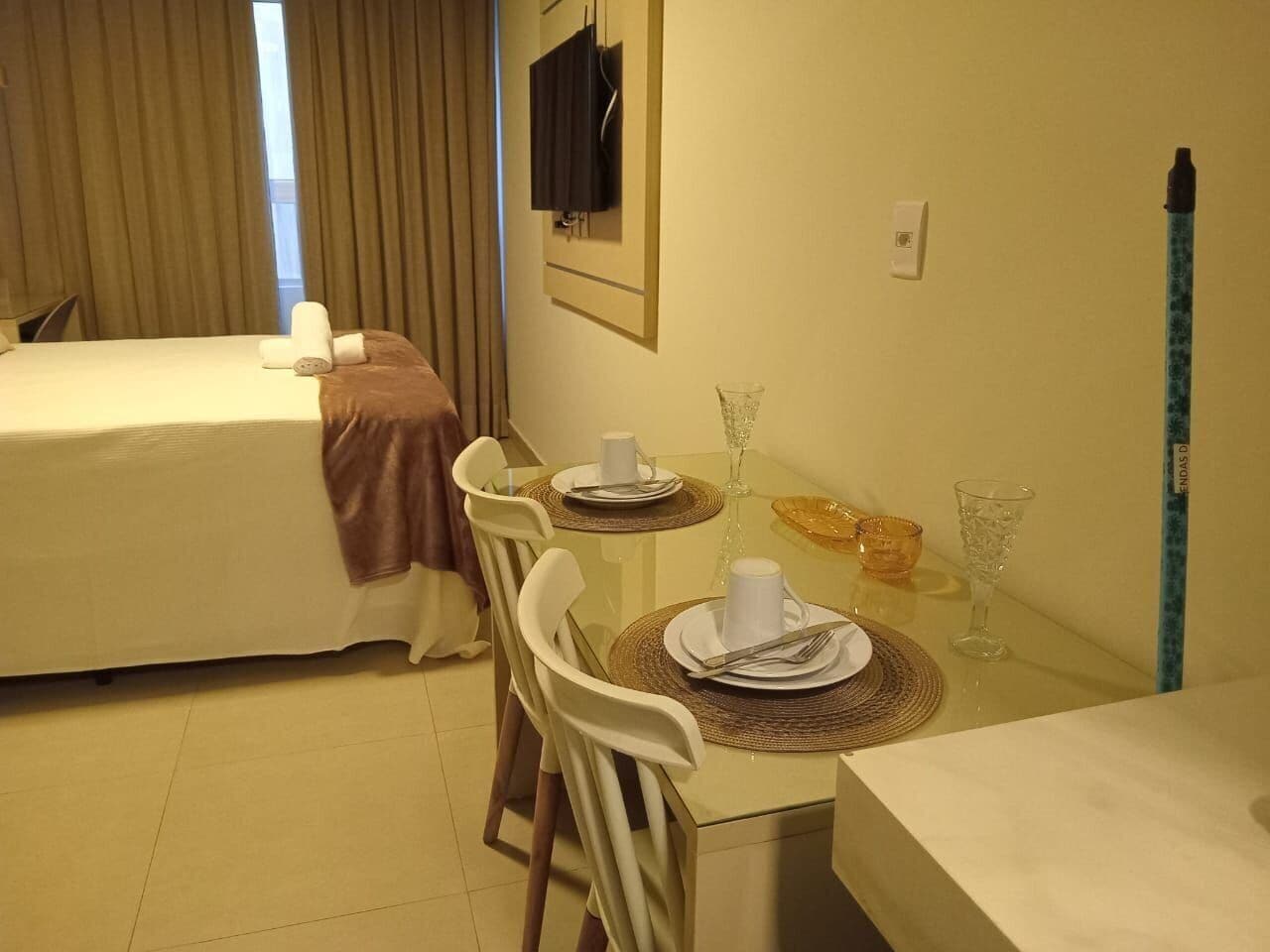 A hotel room with a bed, a dining table set for two, a TV mounted on the wall, and a window with curtains.