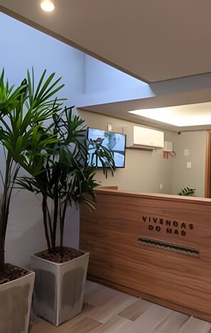 A reception desk with a wooden finish and a sign that reads 'VIVIMDA DO HAUS'. There are potted plants to the left of the desk. - Vivendas do Mar (João Pessoa)