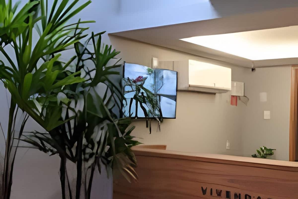 Uma receção com acabamento em madeira e um letreiro com a inscrição 'VIVIMODAL DO NAI'. Há plantas em vasos à esquerda e um quadro emoldurado acima do balcão.