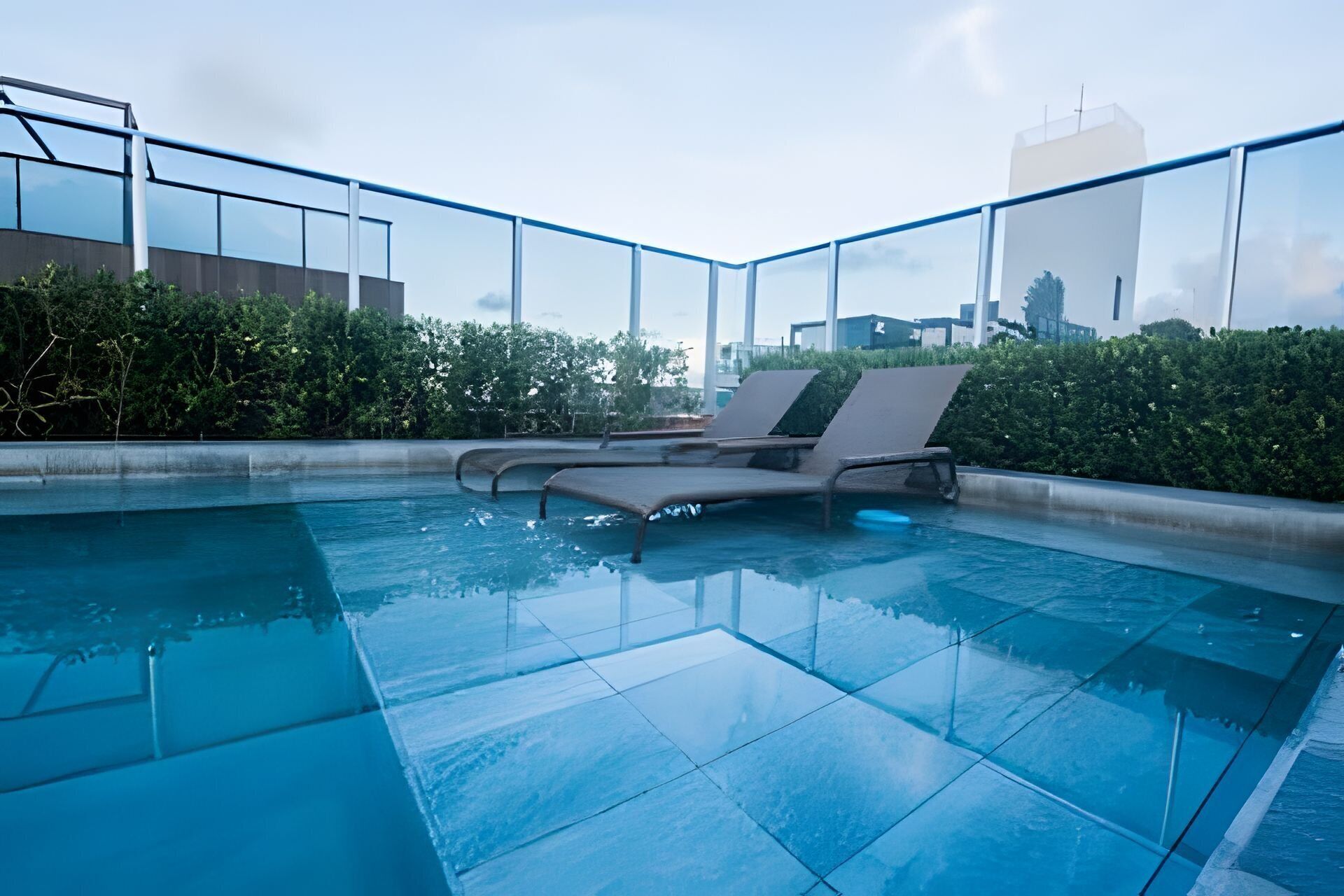 A rooftop pool with clear glass walls, a lounge chair, and a cityscape view.
